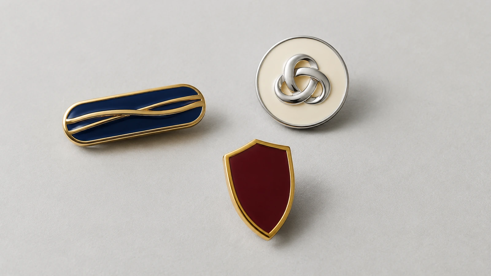 Pins minimalistas em branco com fitas de metal em relevo lisas e um formato de laço entrelaçado que sugerem tipografia, sem nenhum caractere legível