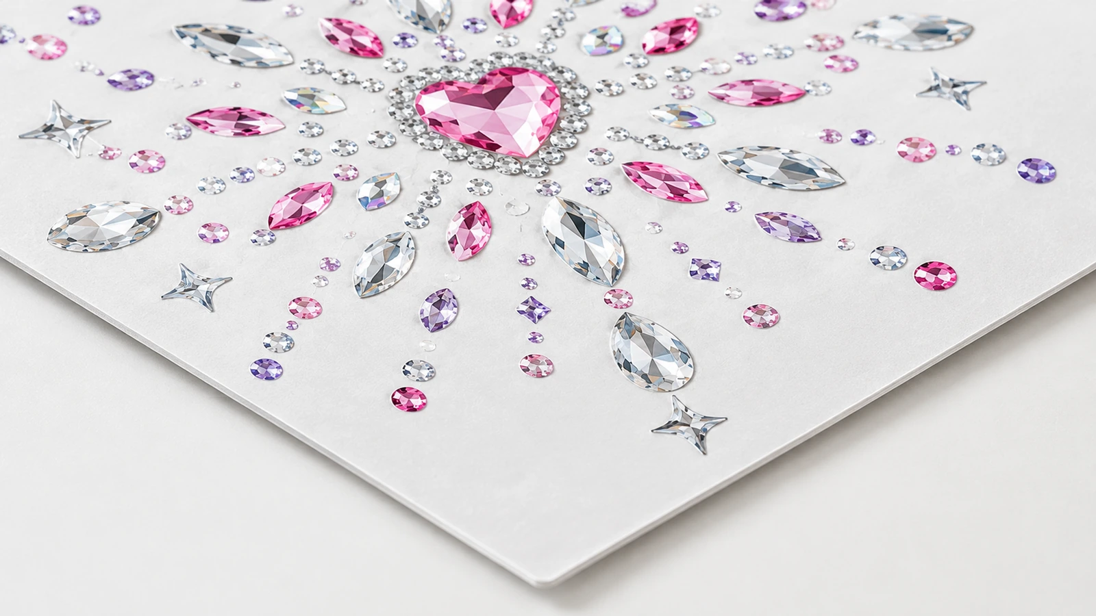 Detalhe ampliado de um motivo de strass rosa com centro em formato de coração e pedras transparentes, rosa e roxas, sobre uma folha branca