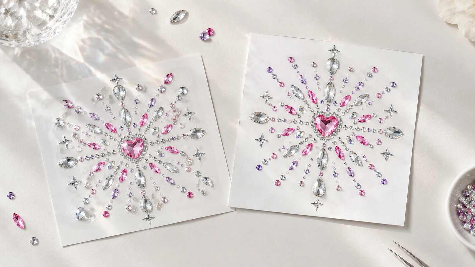 Dois motivos de strass iguais com centro em formato de coração sobre folhas brancas, com pedras rosa, roxas e transparentes